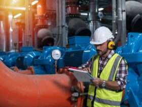 A worker in a hard hat and safety vest checks a clipboard next to a pressure gauge on industrial piping.