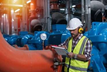 A worker in a hard hat and safety vest checks a clipboard next to a pressure gauge on industrial piping.