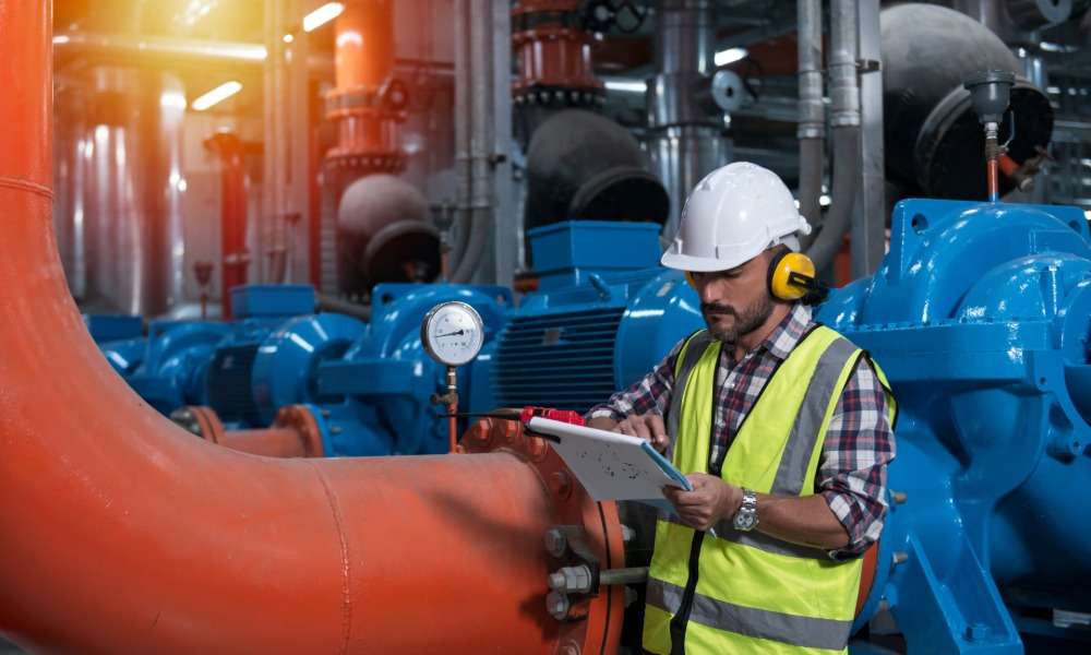 A worker in a hard hat and safety vest checks a clipboard next to a pressure gauge on industrial piping.
