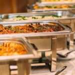 A row of tins containing catered food for a wedding. There are serving spoons on plates next to the tins.