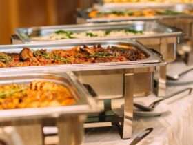 A row of tins containing catered food for a wedding. There are serving spoons on plates next to the tins.