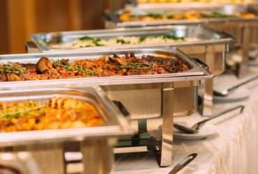 A row of tins containing catered food for a wedding. There are serving spoons on plates next to the tins.