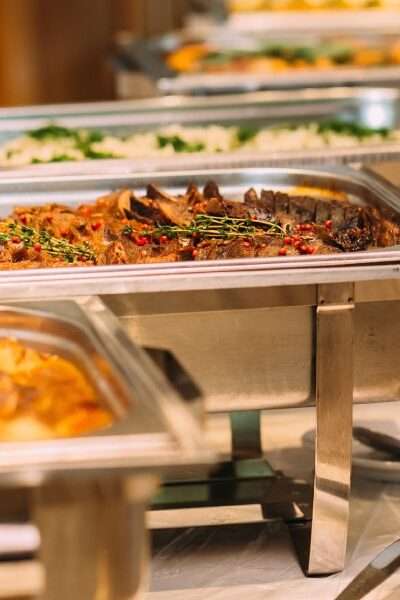 A row of tins containing catered food for a wedding. There are serving spoons on plates next to the tins.