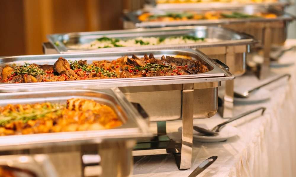 A row of tins containing catered food for a wedding. There are serving spoons on plates next to the tins.