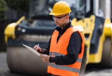 A worker wearing a hard hat, safety vest, and safety glasses while writing on a clipboard. There's a large machine behind him.