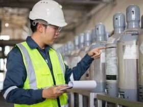 A man wearing a bright yellow safety vest and a white hard hat holds a clipboard and inspects gas cylinders.