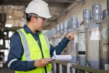 A man wearing a bright yellow safety vest and a white hard hat holds a clipboard and inspects gas cylinders.