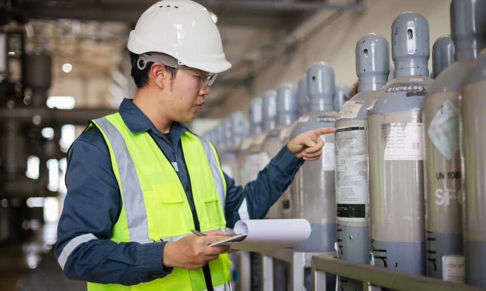 A man wearing a bright yellow safety vest and a white hard hat holds a clipboard and inspects gas cylinders.