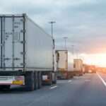 Several semitrucks in a line while stopped in freeway traffic at sunset, billboards in the distance.