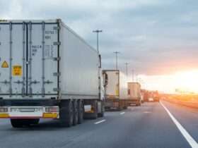 Several semitrucks in a line while stopped in freeway traffic at sunset, billboards in the distance.