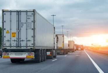Several semitrucks in a line while stopped in freeway traffic at sunset, billboards in the distance.