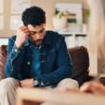 A man sitting in a therapist's office on a couch. He has his hand on his head during a mental health evaluation.