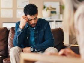 A man sitting in a therapist's office on a couch. He has his hand on his head during a mental health evaluation.
