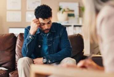 A man sitting in a therapist's office on a couch. He has his hand on his head during a mental health evaluation.
