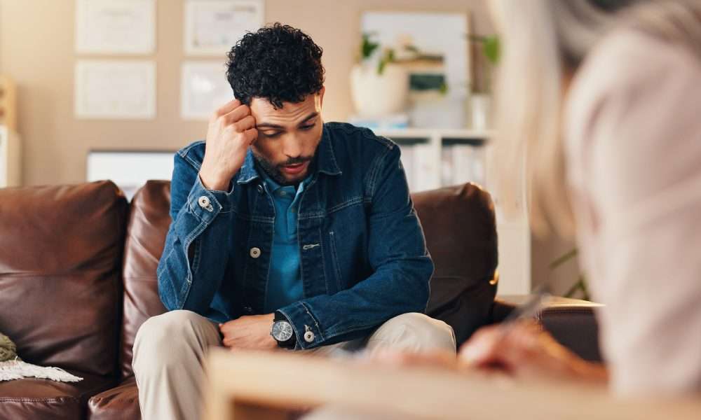 A man sitting in a therapist's office on a couch. He has his hand on his head during a mental health evaluation.
