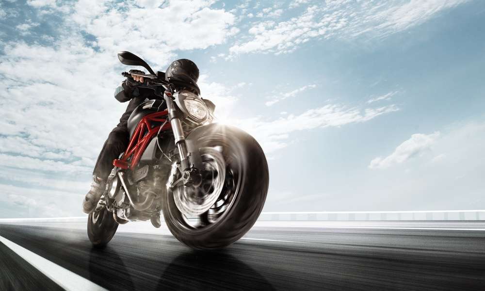 A person wearing a black jacket, black helmet, and black pants is driving a black motorcycle quickly.