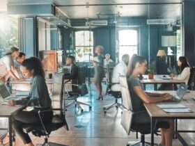An office features people working at various tables and typing on their laptops. A woman walks through, carrying a file.