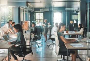 An office features people working at various tables and typing on their laptops. A woman walks through, carrying a file.