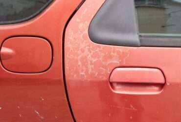 A red car door with faded paint that shows cracks and the underlayer. The damage is on other parts of the car.