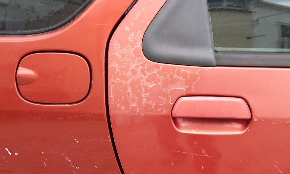 A red car door with faded paint that shows cracks and the underlayer. The damage is on other parts of the car.
