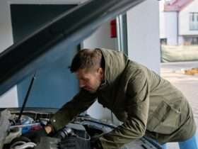 Man in jacket leans over open car hood, inspecting engine components in driveway outside residential home.