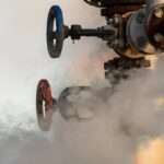 A broken valve on a factory's hydraulic system drips water. White clouds of steam drift from it into the air.