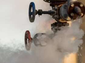 A broken valve on a factory's hydraulic system drips water. White clouds of steam drift from it into the air.