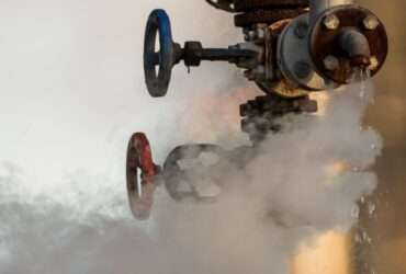A broken valve on a factory's hydraulic system drips water. White clouds of steam drift from it into the air.