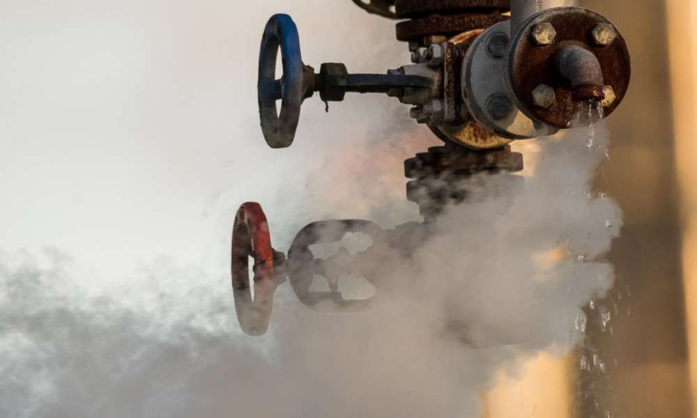 A broken valve on a factory's hydraulic system drips water. White clouds of steam drift from it into the air.