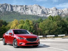 A red car is parked on a paved road with green mountains, trees, and a cloudy daytime sky behind it.
