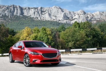 A red car is parked on a paved road with green mountains, trees, and a cloudy daytime sky behind it.
