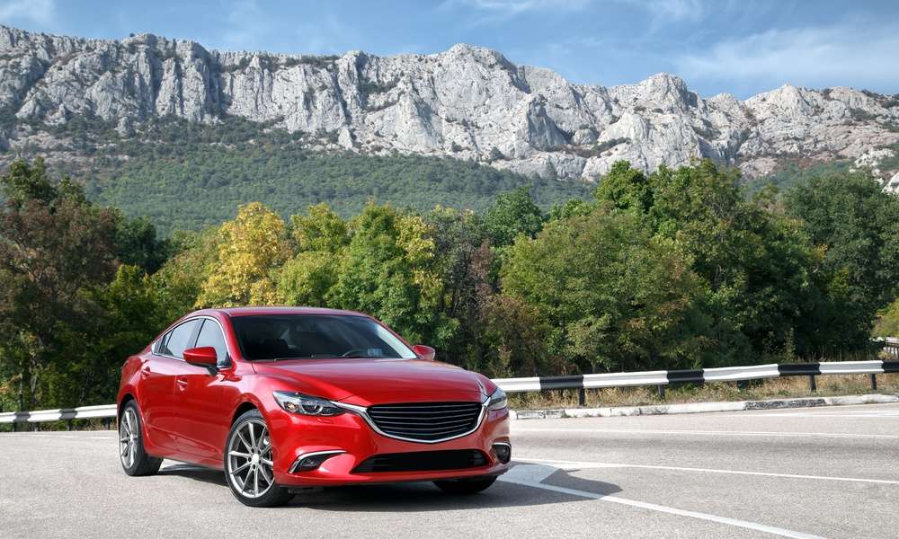 A red car is parked on a paved road with green mountains, trees, and a cloudy daytime sky behind it.