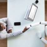 An aerial view of a interviewee shaking the hand of a robot interviewer across a rectangular white table.