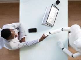 An aerial view of a interviewee shaking the hand of a robot interviewer across a rectangular white table.