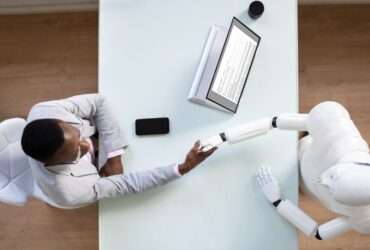 An aerial view of a interviewee shaking the hand of a robot interviewer across a rectangular white table.