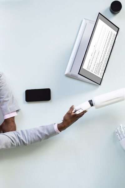 An aerial view of a interviewee shaking the hand of a robot interviewer across a rectangular white table.