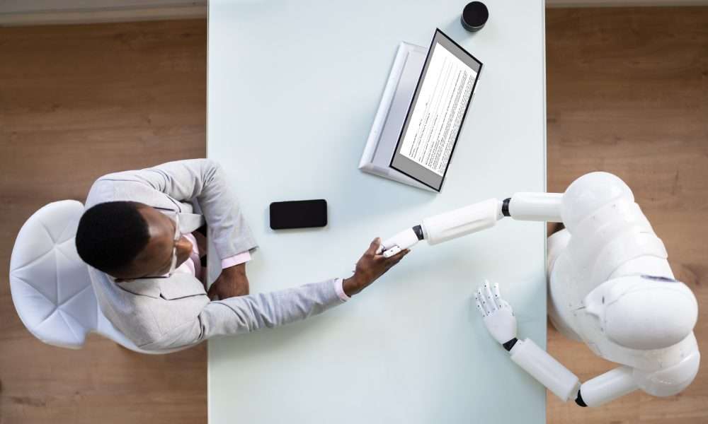 An aerial view of a interviewee shaking the hand of a robot interviewer across a rectangular white table.