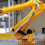 A bright yellow industrial robotic arm lifting a metal gripper inside a factory, surrounded by machinery and safety fencing.