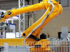 A bright yellow industrial robotic arm lifting a metal gripper inside a factory, surrounded by machinery and safety fencing.
