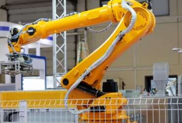 A bright yellow industrial robotic arm lifting a metal gripper inside a factory, surrounded by machinery and safety fencing.