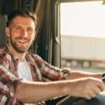 A trucker with a wide grin sits in the cab of a semitruck, both of his hands on the steering wheel.