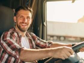 A trucker with a wide grin sits in the cab of a semitruck, both of his hands on the steering wheel.