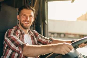 A trucker with a wide grin sits in the cab of a semitruck, both of his hands on the steering wheel.