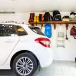 Interior garage with white hatchback, neatly arranged shelves, tools, vacuum, ladder, and storage items on wall.