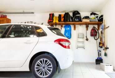 Interior garage with white hatchback, neatly arranged shelves, tools, vacuum, ladder, and storage items on wall.