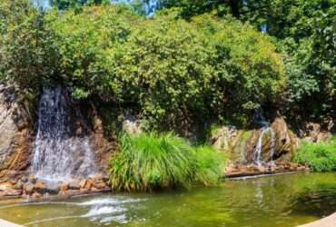 Water flows from tall rocks into an artificial lake, creating a waterfall. Trees and shrubs surround the waterfalls.