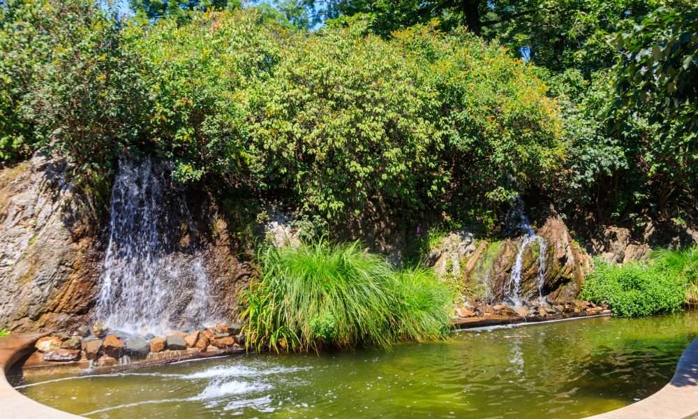Water flows from tall rocks into an artificial lake, creating a waterfall. Trees and shrubs surround the waterfalls.