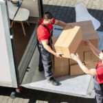 Two men, both of whom are wearing red shirts, are working together to take boxes out of the back of a moving truck.