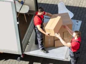 Two men, both of whom are wearing red shirts, are working together to take boxes out of the back of a moving truck.