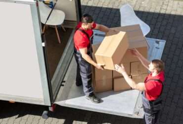 Two men, both of whom are wearing red shirts, are working together to take boxes out of the back of a moving truck.